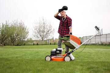 Tired man in casual clothes holds bag with cut grass of lawn mower after mowing lawn with at own backyard. Husband takes care of garden. Modern gasoline garden equipment. Landscaping work at home