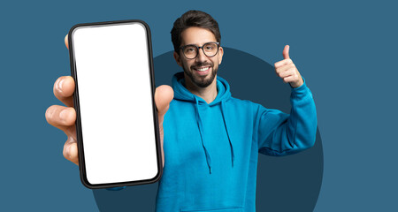 A young man smiling and giving a thumbs up is holding a blank smartphone in front of him. He wears a blue hoodie and stands against a plain background that enhances his cheerful expression.