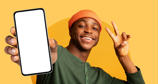 Young man wearing an orange beanie holds up a smartphone with a blank screen and smiles joyfully while making a peace sign. The image features a vibrant yellow backdrop.