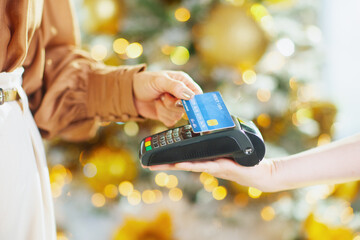 A close-up shows womans hand making contactless credit card payment at point-of-sale terminal. Blurred festive lights in background suggest modern holiday shopping convenience and quick transaction
