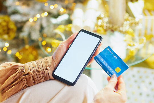 Close-up of hands using a smartphone and credit card for holiday online purchases. The soft, festive background hints at the convenience of digital payment during the season.