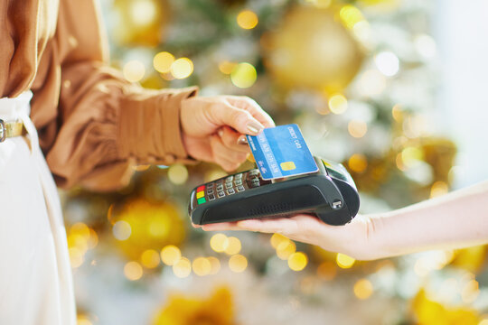 A close-up shows womans hand making contactless credit card payment at point-of-sale terminal. Blurred festive lights in background suggest modern holiday shopping convenience and quick transaction