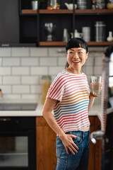 Smiling Japanese woman holding a glass of water in modern kitchen. Cheerful Asian woman promoting hydration, wellness and healthy lifestyle with natural light and relaxed mood.