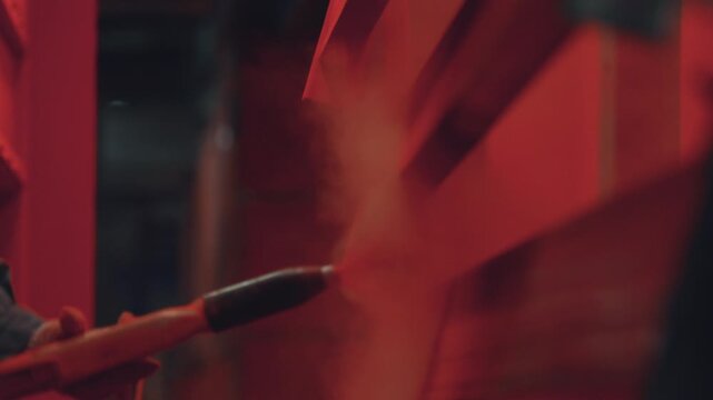Factory worker wearing protective gloves using a spray gun to apply red powder coating to a metal structure in an industrial workshop, showcasing the manufacturing and finishing process
