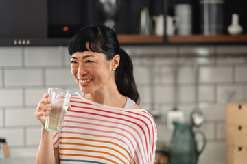 Smiling Japanese woman holding a glass of water in modern kitchen. Cheerful Asian woman promoting hydration, wellness and healthy lifestyle with natural light and relaxed mood.