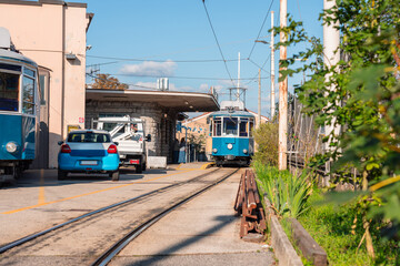 Naklejka premium Historic blue Opicina tram arriving at a small station in Opicina, Italy. The vintage streetcar passes along narrow tracks near parked cars, with sunlight casting warm tones on the scene.