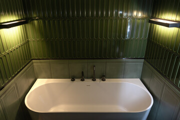 Bathtub with dark fixtures amid green tiles and soft light. Evening relax.