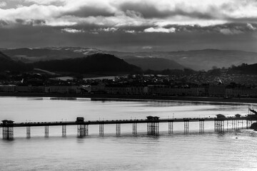 Monochrome view of Llandudno Pier stretching into the sea with calm water, seafront buildings, and hills in the background under dramatic clouds
