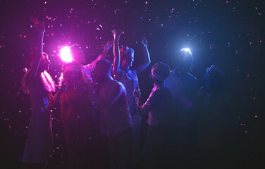 A group of friends enjoys a festive moment, dancing and celebrating together surrounded by colorful lights and confetti at a nighttime gathering.
