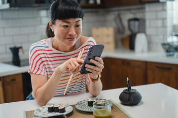Cheerful Japanese woman eating sushi with chopsticks while looking at smartphone in kitchen. Happy Asian woman enjoying meal, technology and modern home lifestyle