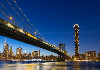 Fototapeta premium View of New York City and the Brooklyn Bridge with city lights at night near the water, at night.