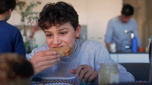 Boy eating slice of bread with spread during breakfast at home kitchen table with family in background