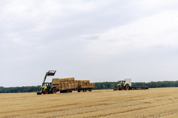 Obraz premium Tractors Transporting Straw Bales Across Fields Under Overcast Skies in Rural Landscape
