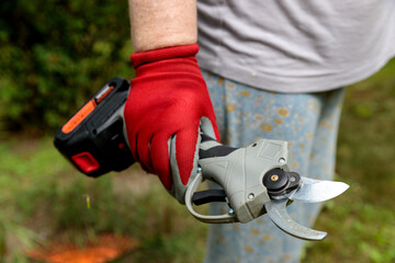 Powerful Gardening Tool: A Close-Up View of an Electric Pruner Held by a Gardener in Red Gloves Ready for Pruning and Trimming Plants in a Green Garden Environment.