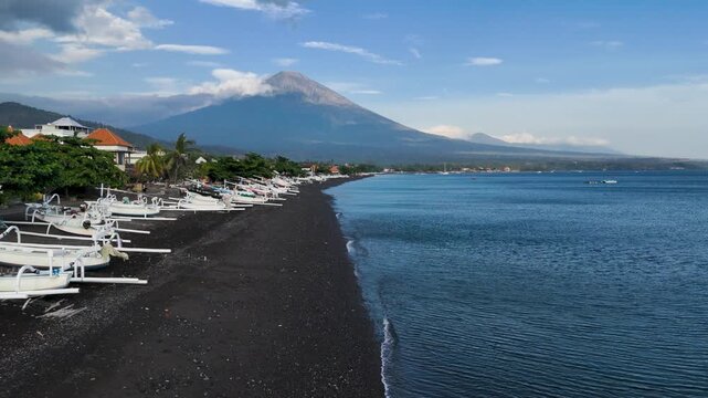 Drone aerial view of Amed beach, Bali, lined up with fishing boats with black sand and calm sea water, Mount Agung in the background.