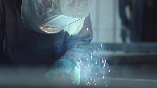 Close up view of a factory worker in a protective mask welding steel structures, creating bright flashes of light, sparks and smoke during the metalworking process in a workshop