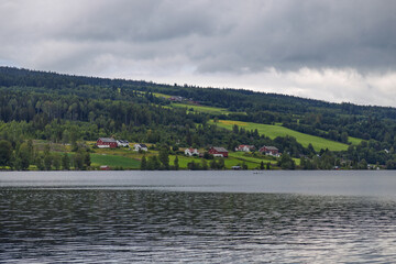 Serene Lakeside Landscape with Traditional Homes Nestled Among Forested Hills Under Cloudy Skies