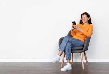 Cheerful positive attractive young indian woman wearing casual outfit sitting in armchair at home,...