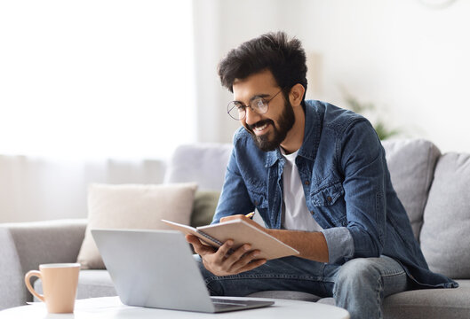 Distance Learning. Young Indian Man Study With Laptop At Home And Taking Notes, Millennial Eastern Guy In Eyeglasses Watching Online Webinar, Sitting On Couch With Computer, Enjoying Remote Education - Powered by Adobe