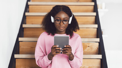 African American Student Lady Using Digital Tablet Learning Online Or Watching Movie Wearing Wireless Headphones Sitting On Steps Of Staircase At Home. E-Learning, Gadgets And Technology Concept