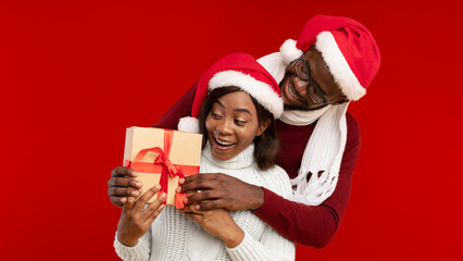 Christmas Gifts. African American Husband Surprising Excited Wife Giving Wrapped Present Box Hugging Standing Over Yellow Studio Background, Wearing Santa Hats. Merry Xmas Concept