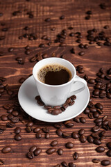 A cup of dark roasted coffee on a white saucer, surrounded by coffee beans on a brown wooden surface