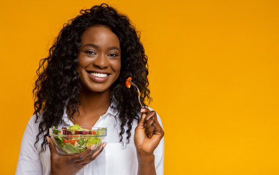 A joyful woman holds a bowl of mixed salad, smiling brightly while using a fork to pick up a piece of tomato. The vibrant yellow background enhances the lively atmosphere.
