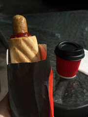 French hot dog in paper wrapper in hand and cup of coffee on glass table. Concept for quick snacks and takeaway food. Street cafe. 
