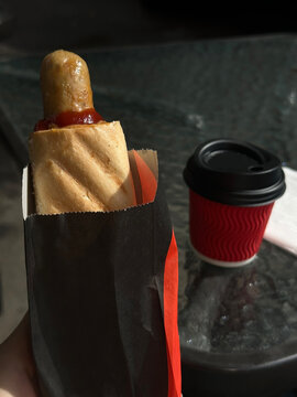 French hot dog in paper wrapper in hand and cup of coffee on glass table. Concept for quick snacks and takeaway food. Street cafe. 