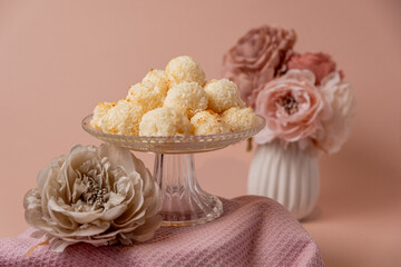 Delicious coconut dessert balls arranged elegantly on a glass cake stand, surrounded by soft pastel flowers and a textured fabric, creating a charming and inviting atmosphere