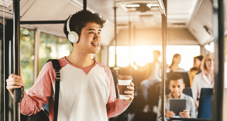 A young man stands inside a bus, wearing headphones and smiling. He holds a coffee cup in one hand while gripping a handrail with the other.