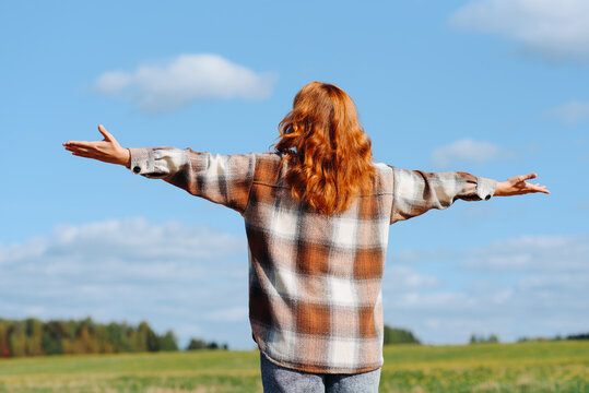 young beautiful redhead caucasian woman standing on country road, stretching hands to sky, beige plaid shirt, traveling alone, sunny summer day, blue sky, green field and trees, beautiful landscape