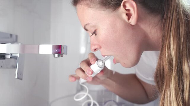 A young woman uses a water flosser to clean her teeth in the bathroom in the morning. Concept: morning routine and oral hygiene at home.