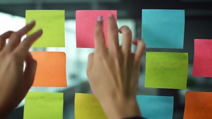 Woman's hand places sticky notes of various colors on transparent glass in a modern workspace - Powered by Adobe