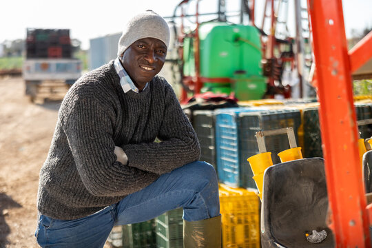 Smiling adult african american farmer standing outdoors leaning on farm machine on sunny spring day.. - Powered by Adobe