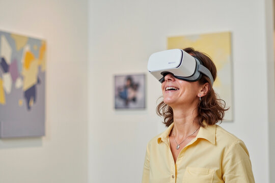 Caucasian middle aged woman wearing virtual reality headset standing indoors smiling and experiencing immersive digital content with abstract paintings visible on wall in background