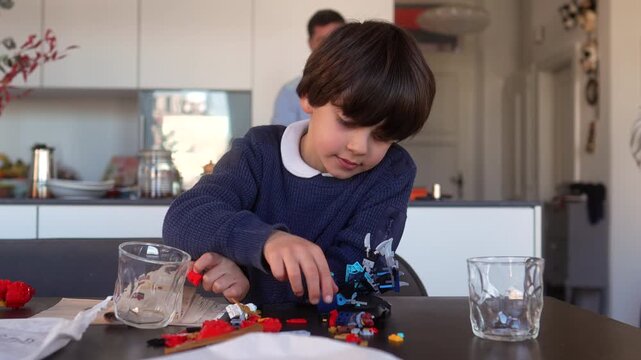 Young boy concentrating on building colorful plastic toy blocks while sitting at kitchen table with father preparing drink in background at modern family home