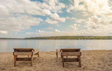 Wooden sun loungers on sandy beach by calm lake under blue sky with clouds, peaceful summer landscape