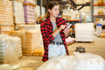 Young european woman warehouse worker using smartphone to check quantity of goods.
