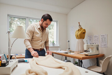 Caucasian young adult man working with fabric on table in sewing studio, using hands to measure material, sewing machine and dress form visible in background, fashion design process