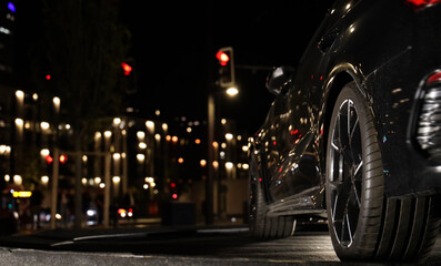 Close-up of black rear wheel with sporty low profile  tire. Five spoke silver rim, visible brake caliper and disc, glossy black car body in background Light bokeh illuminated  Night scene city life