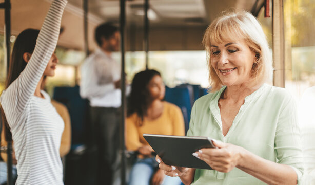 A woman smiles while using a tablet on a bus. She is surrounded by other passengers, including a person standing and others sitting. - Powered by Adobe
