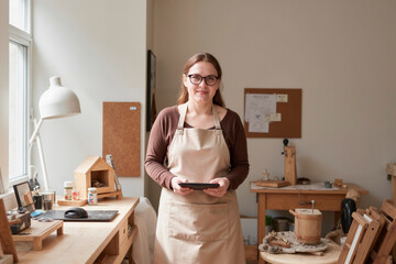 Portrait of Caucasian young adult woman wearing glasses standing in workshop holding digital tablet, smiling at camera, surrounded by woodworking tools and handmade crafts