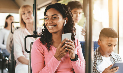 A happy woman in a pink long-sleeve shirt holds her smartphone on a bus. She wears earbuds and enjoys the moment, surrounded by other passengers engaged in their devices.
