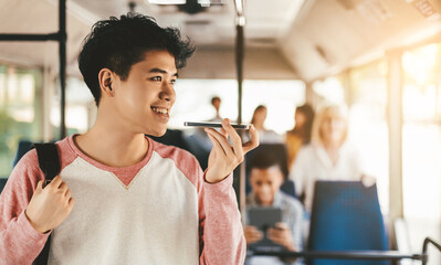 A young man smiles while using his phone's voice assistant on a bus. Other passengers are seated, some engaged with devices, creating a relaxed atmosphere during the commute.