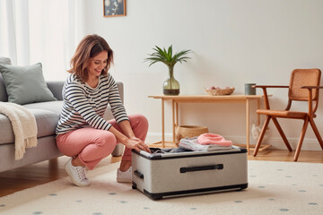 Caucasian young adult woman kneeling on carpet packing suitcase with folded clothes, smiling while preparing for travel in modern living room with minimal decor visible in background
