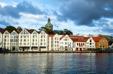 Waterfront view of Stavanger harbor in Norway, with colorful traditional wooden houses, boats, and calm water under dramatic clouds at sunset.