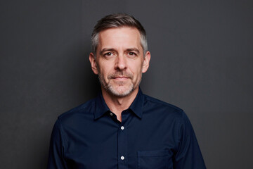 Obraz premium Portrait of middle aged Caucasian man looking into camera with slight smile, short gray hair and trimmed beard, standing against plain dark background, neutral expression