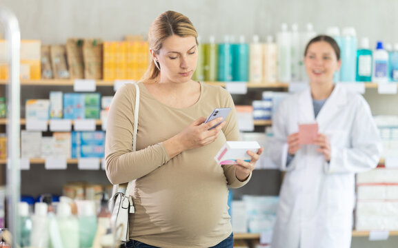 Focused pregnant woman scanning QR code on package of medicine with smartphone, checking potential side effects while pharmacist waiting to assist with professional advice standing in background.. - Powered by Adobe