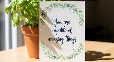 A motivational card with the text "You are capable of amazing things" surrounded by a floral wreath, placed next to a potted basil plant.